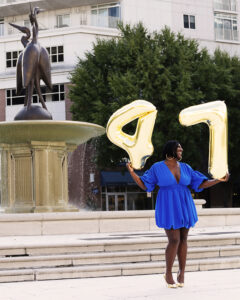 A joyful woman holding birthday balloons poses in front of the fountains at Town Center, Virginia Beach, smiling as the water cascades behind her in Virginia Beach Town Center.